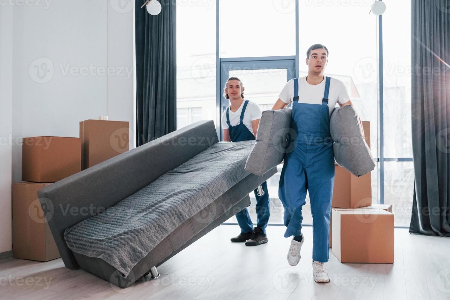 schweres Sofa tragen. Zwei junge Umzugshelfer in blauer Uniform arbeiten drinnen im Raum foto