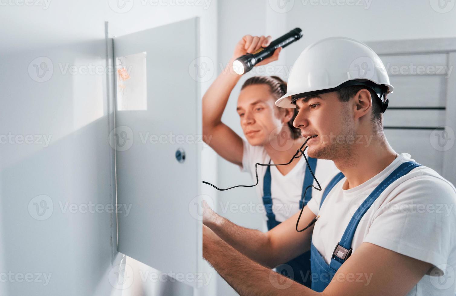 zwei junge männliche elektriker arbeiten drinnen zusammen. mit Taschenlampe foto