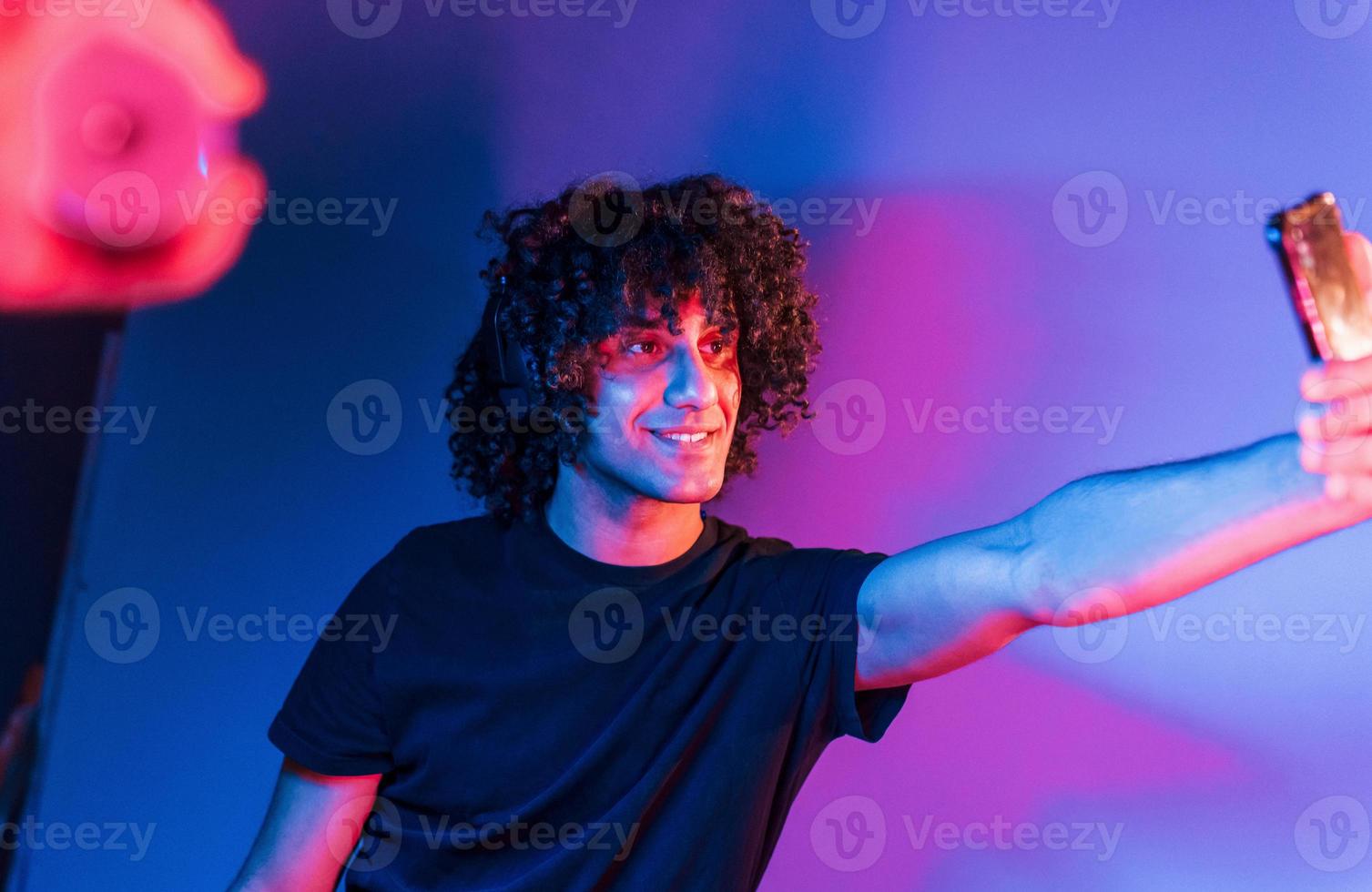 macht ein Selfie. junger schöner mann mit lockigem haar ist drinnen im studio mit neonbeleuchtung foto