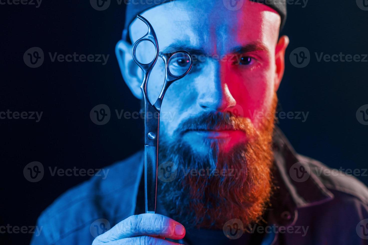 mit Schere. junger bärtiger friseur in kappe, der im studio mit neonbeleuchtung steht foto