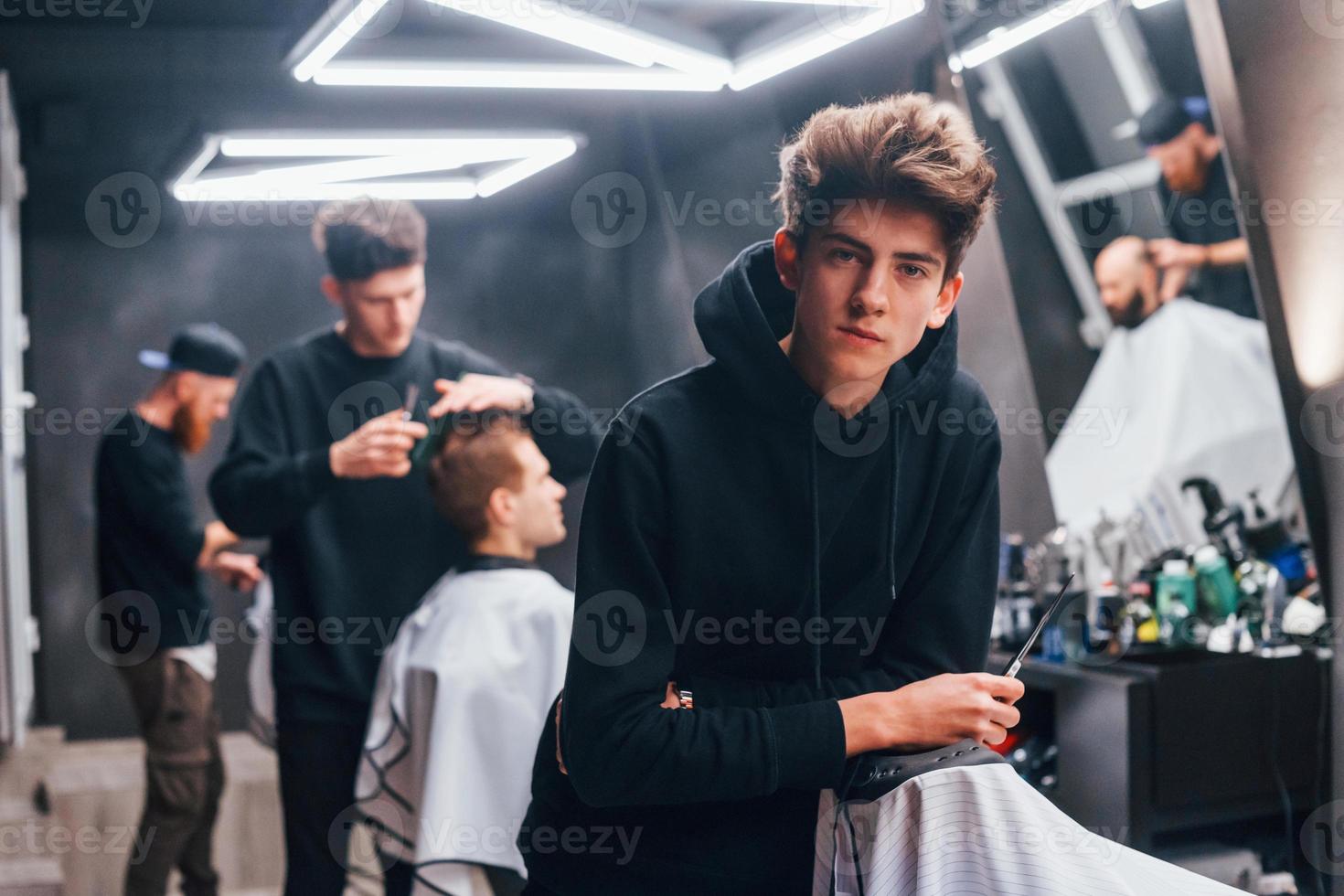 junger friseur in schwarzer kleidung, der im friseursalon steht. Arbeiter und Kunden hinter sich foto