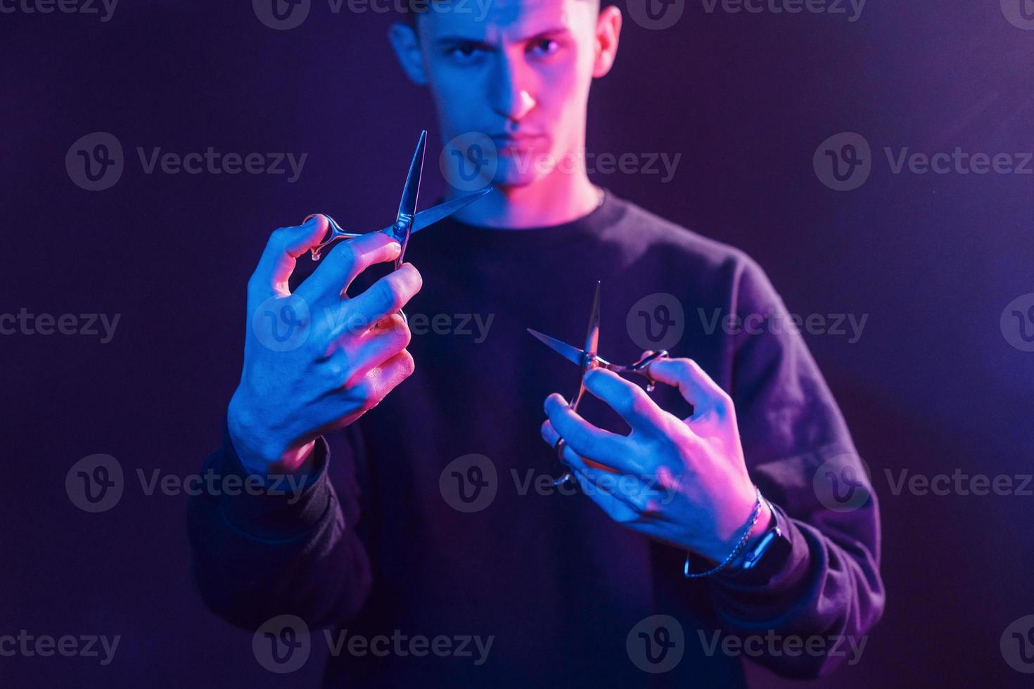 hält eine Schere. junger friseur mit arbeitsgeräten, der im studio mit neonbeleuchtung steht foto