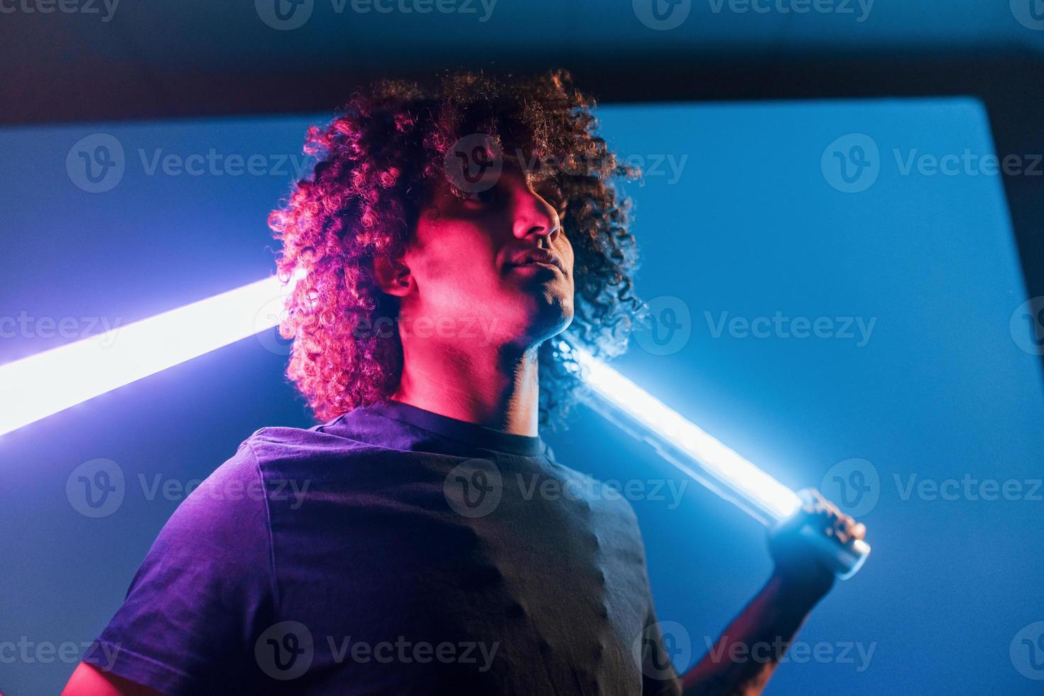 hält Beleuchtungsgeräte. junger schöner mann mit lockigem haar ist drinnen im studio mit neonbeleuchtung foto