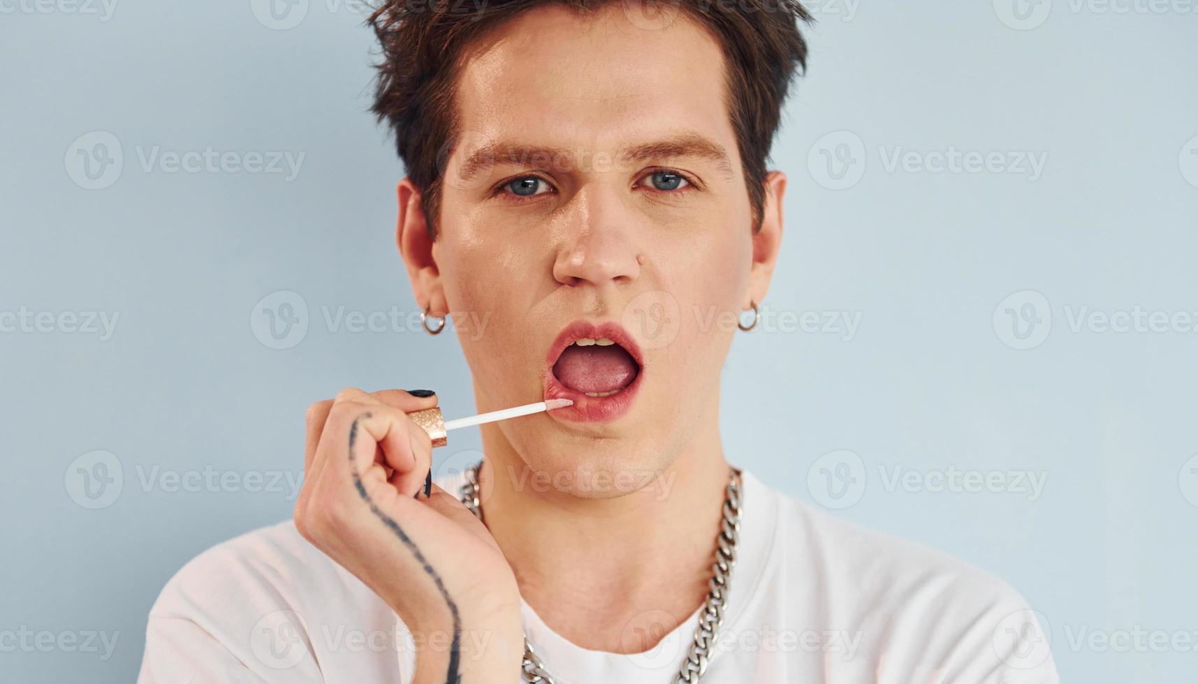 junger schwuler mann steht im studio und posiert für eine kamera. malt Lippen mit Lippenstift foto