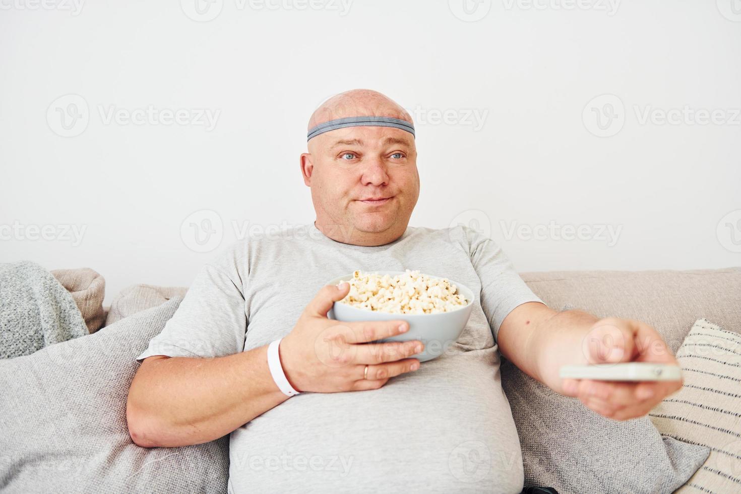 sitzt mit Popcorn auf dem Sofa. lustiger übergewichtiger mann in lässiger kleidung ist drinnen zu hause foto