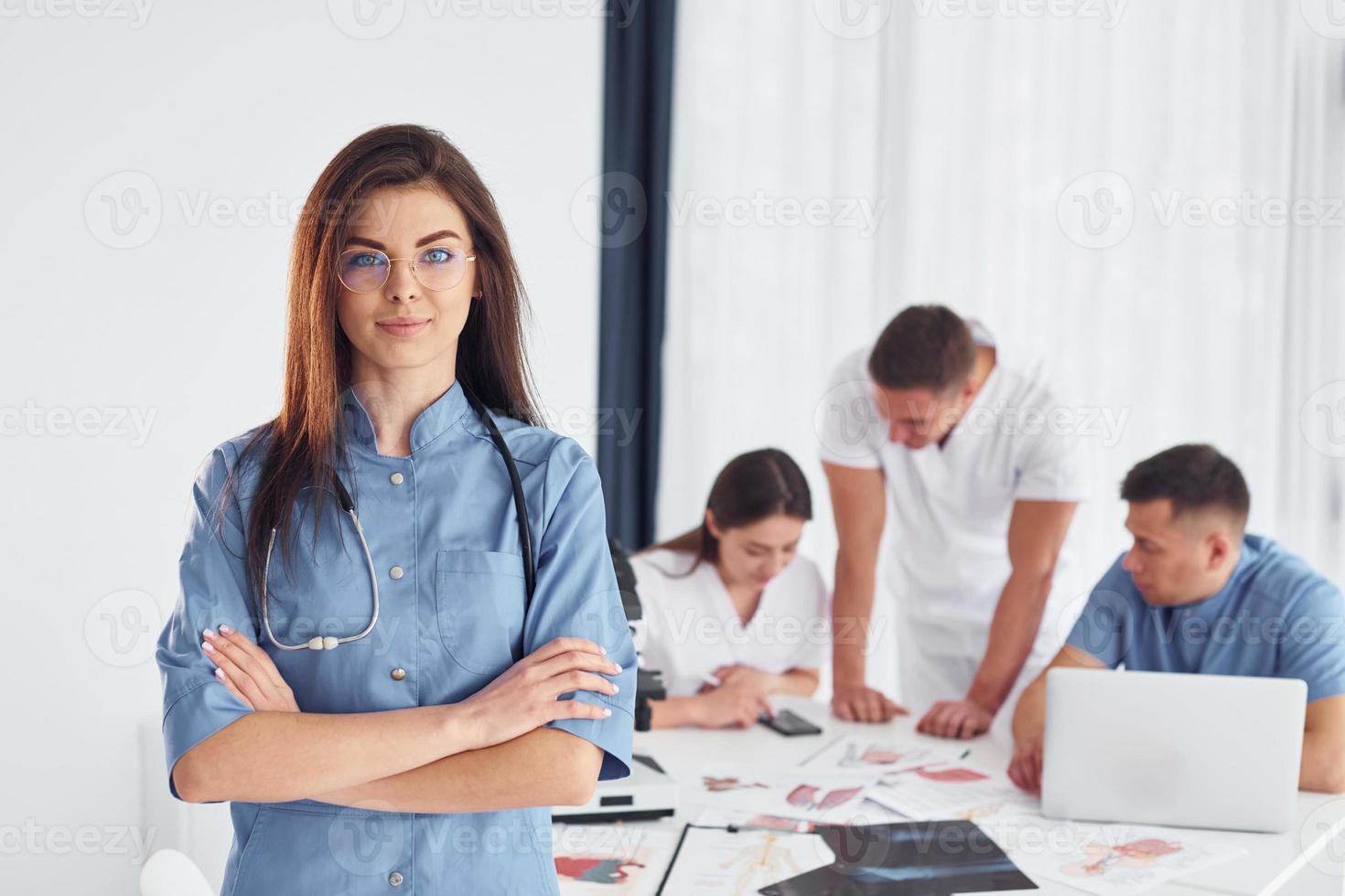 Frau, die gegen eine Gruppe junger Ärzte steht, die im modernen Büro zusammenarbeiten foto