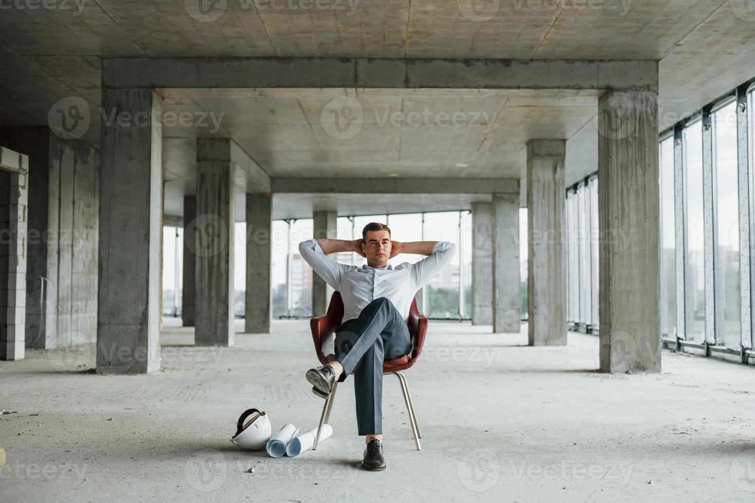 sitzt auf dem Stuhl. junger mann in formeller kleidung arbeitet drinnen am bau foto