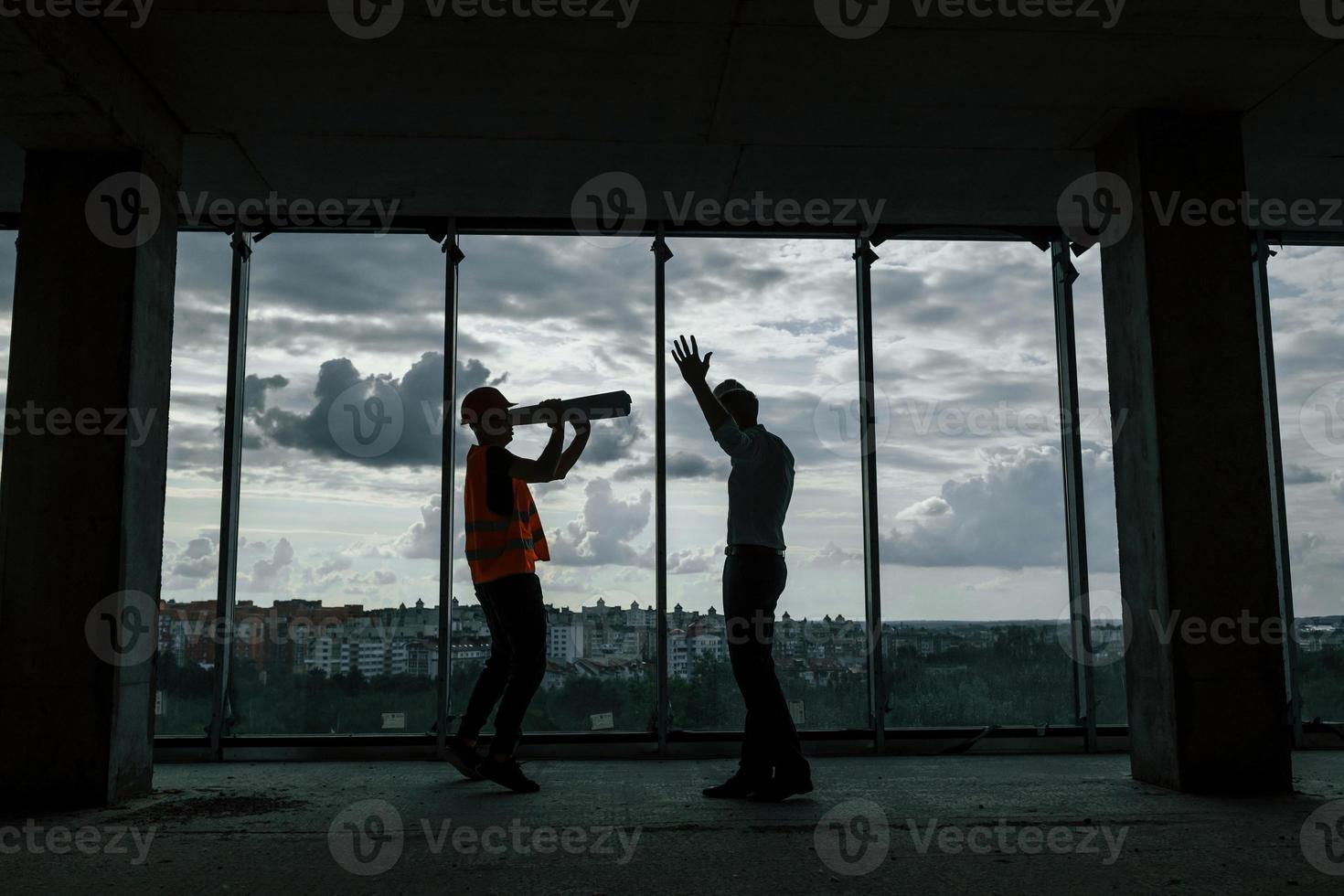 stehend gegen großes Fenster. mann im anzug und handwerker in oranger schutzkleidung arbeitet am bau foto