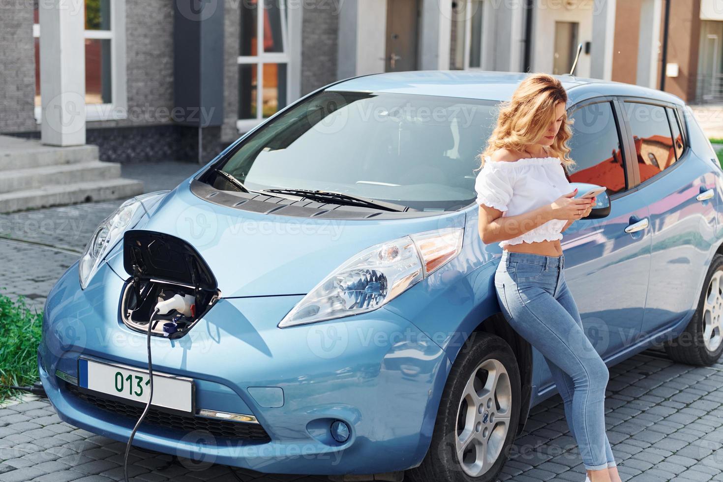 neben dem Auto stehen. Junge Frau in Freizeitkleidung mit ihrem Elektromobil tagsüber im Freien foto