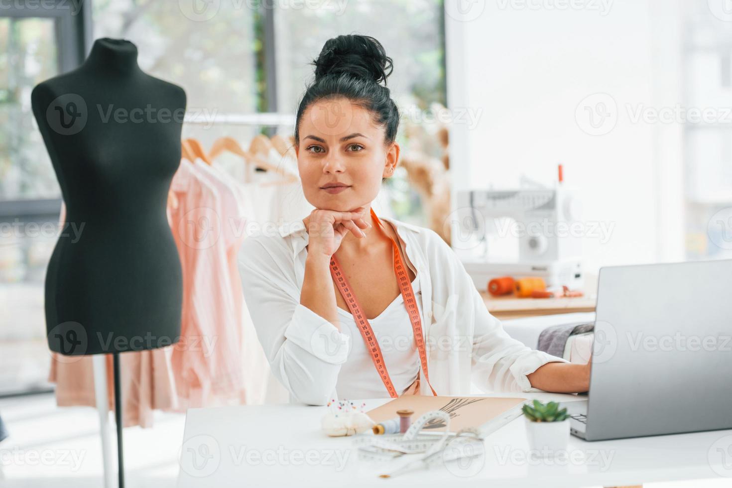 Arbeiten mit Laptop. Näherin ist in ihrem Büro, das mit verschiedenen Kleidern ist foto