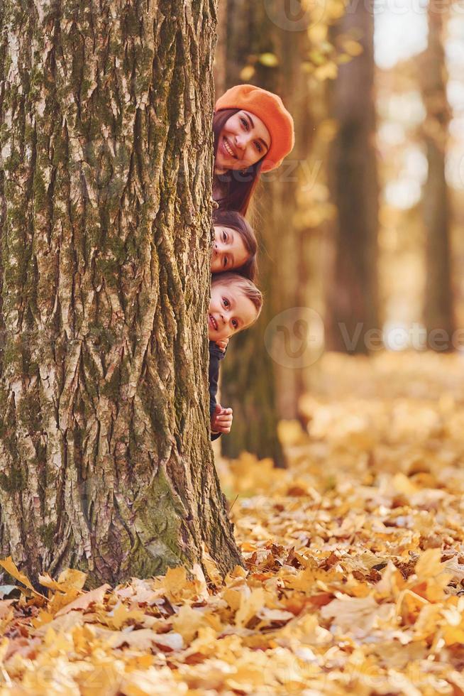 versteckt sich hinter Baum. mutter mit ihrem kleinen sohn und ihrer tochter hat spaß im herbstpark foto