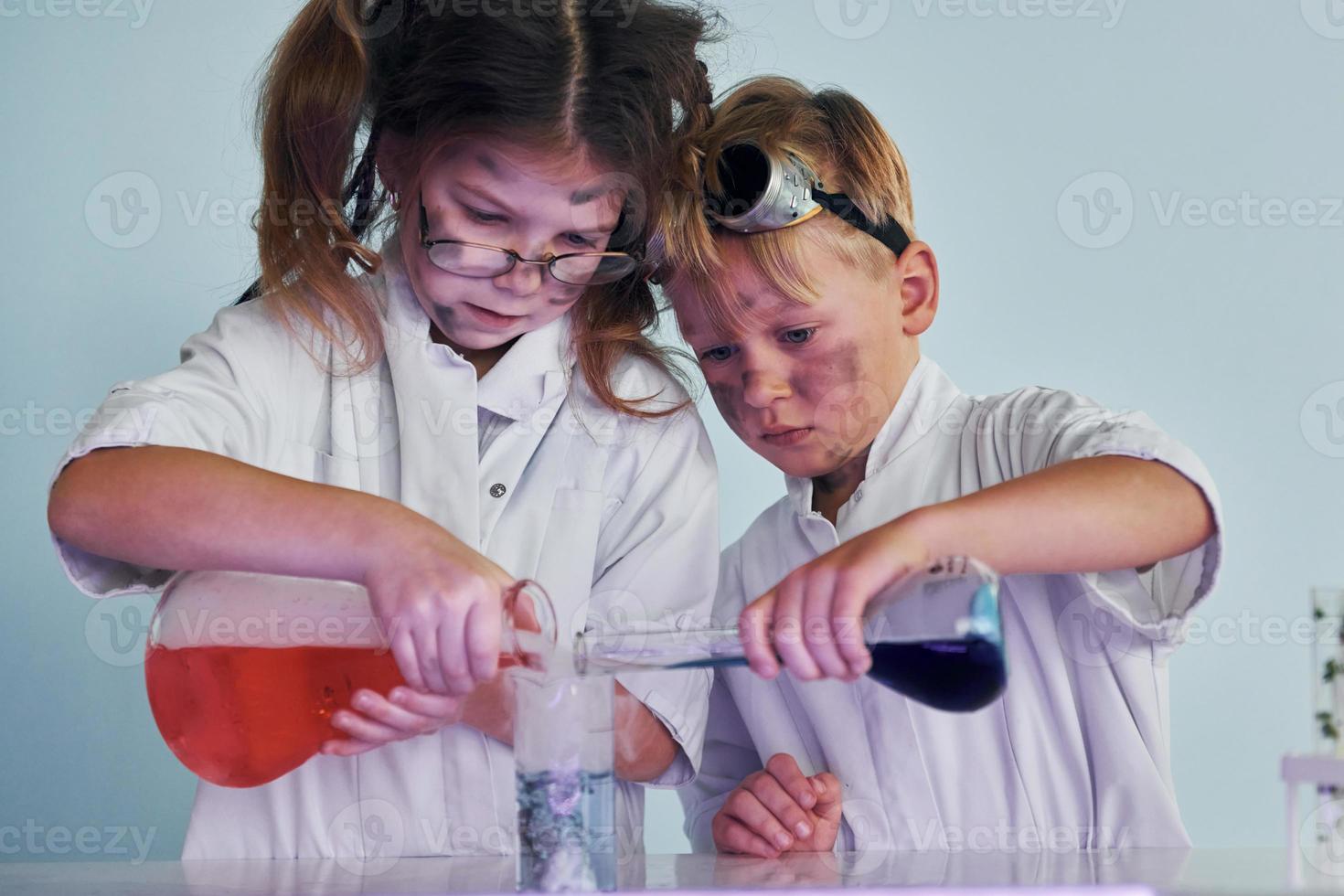 Kleines Mädchen und Junge in weißen Mänteln spielen einen Wissenschaftler im Labor, indem sie Geräte verwenden foto