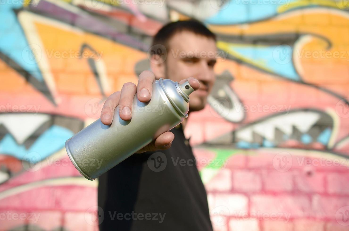 Der junge Graffiti-Künstler zielt mit seiner Sprühdose auf den Hintergrund bunter Graffiti in Rosatönen an der Ziegelwand. Street Art und zeitgenössischer Malprozess foto