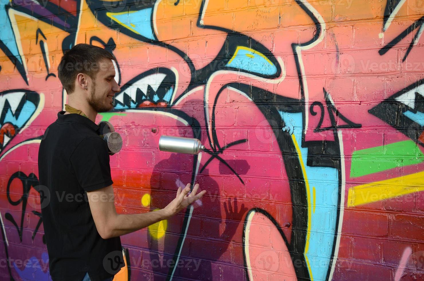 Der junge Graffiti-Künstler mit Gasmaske am Hals wirft seine Sprühdose gegen bunte rosafarbene Graffiti an der Ziegelwand. Street Art und zeitgenössischer Malprozess foto