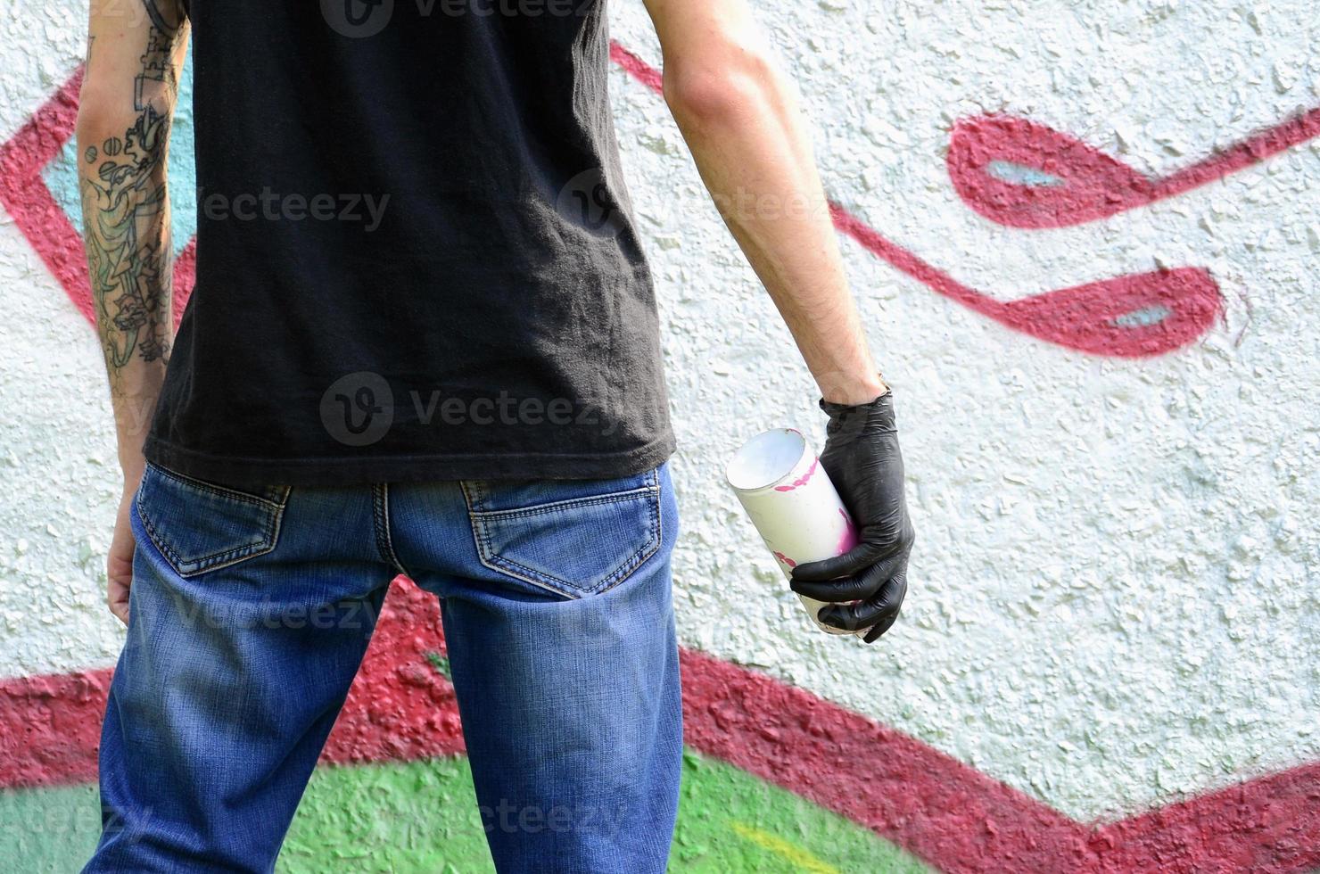 Ein junger Hooligan mit einer Spraydose steht an einer Betonwand mit Graffiti-Gemälden. illegales vandalismuskonzept. Straßenkunst foto