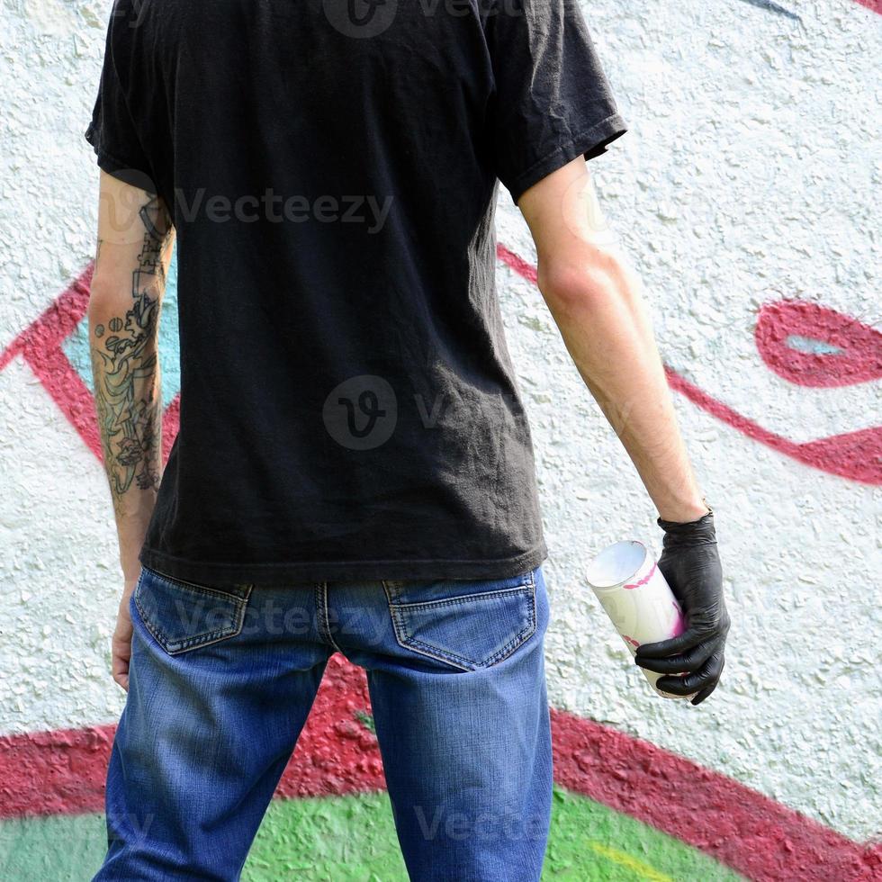 Ein junger Hooligan mit einer Spraydose steht an einer Betonwand mit Graffiti-Gemälden. illegales vandalismuskonzept. Straßenkunst foto