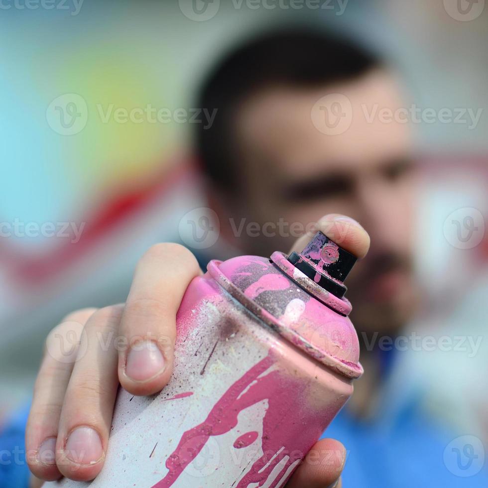 Ein junger Graffiti-Künstler in einer blauen Jacke hält vor dem Hintergrund einer farbigen Graffiti-Zeichnung eine Farbdose vor sich. Straßenkunst- und Vandalismuskonzept foto