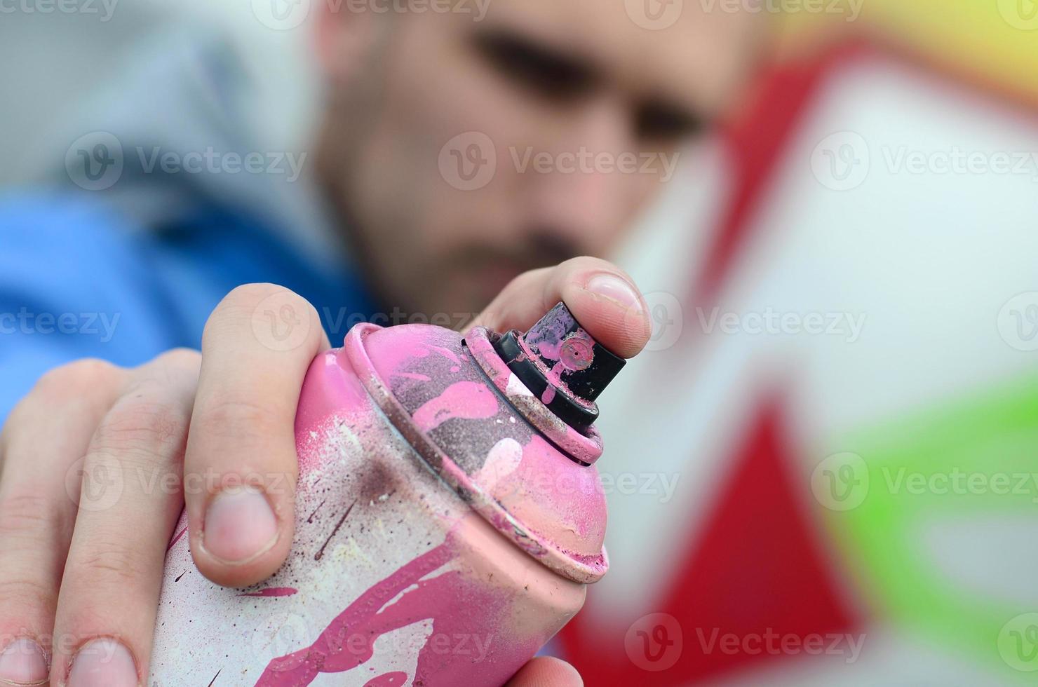 Ein junger Graffiti-Künstler in einer blauen Jacke hält vor dem Hintergrund einer farbigen Graffiti-Zeichnung eine Farbdose vor sich. Straßenkunst- und Vandalismuskonzept foto