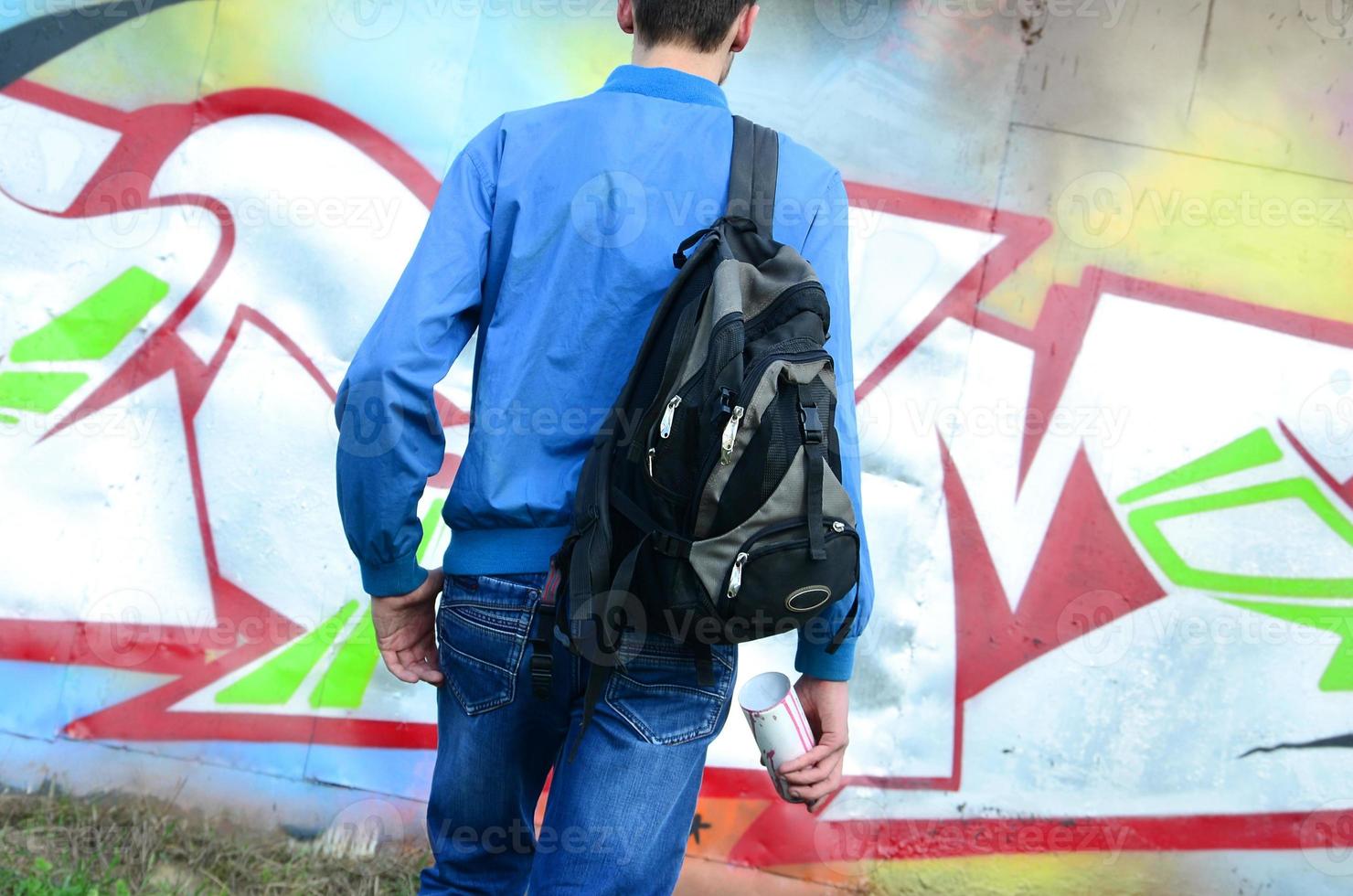Ein junger Graffiti-Künstler mit einer schwarzen Tasche betrachtet die Wand mit seinem Graffiti an einer Wand. Street-Art-Konzept foto