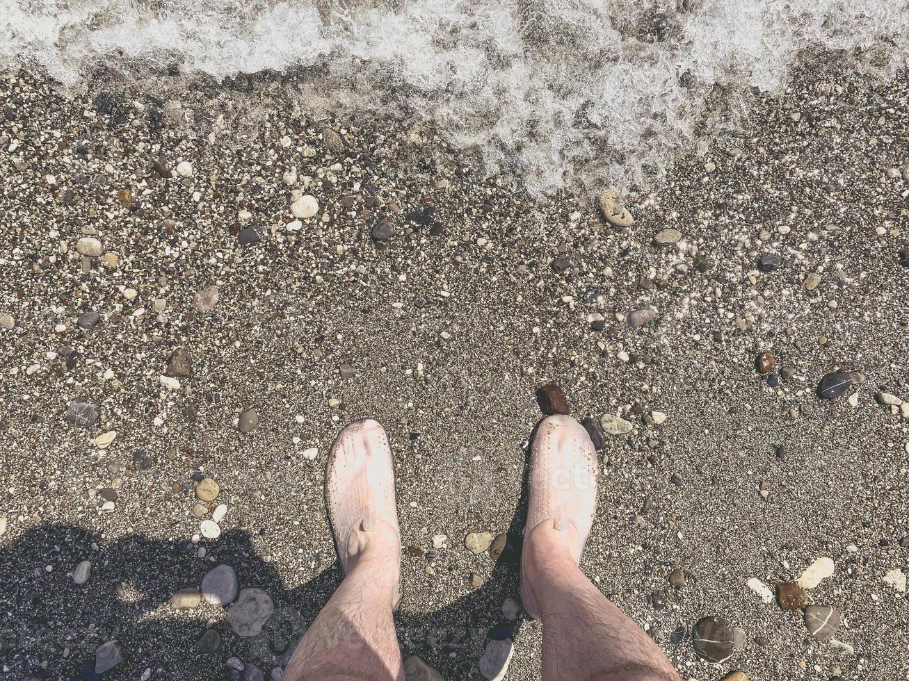 Kiesstrand mit Steinen. ein typ in schuhen steht am ufer. Rest der Touristen auf dem Meer. Füße, die von den Wellen des Meeres gewaschen werden foto