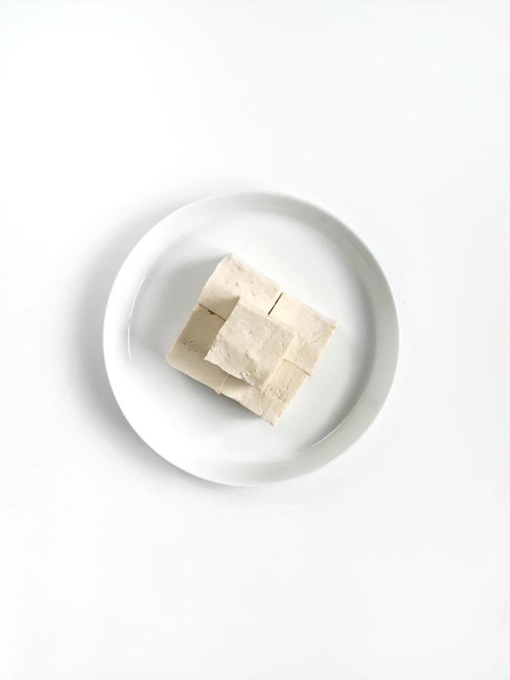 Weißer und glatter Tofu, der in kleinere Stücke geschnitten wird, die auf einer weißen Keramikplatte isoliert auf weißem Hintergrund platziert werden. Ansicht von oben foto
