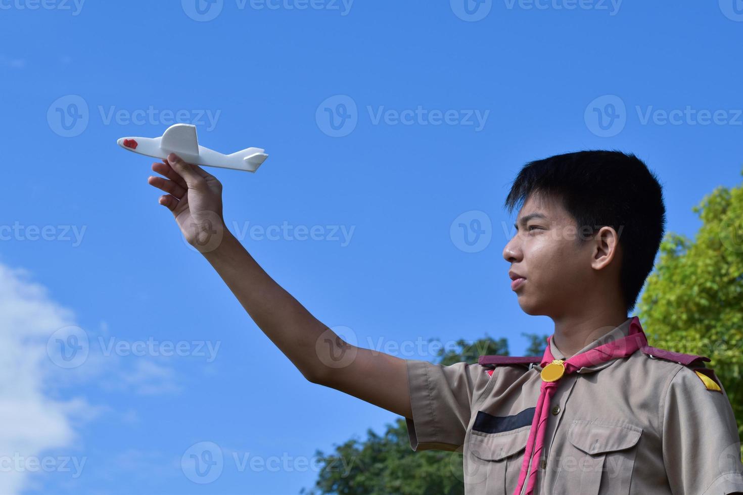 asiatischer pfadfinder hält weißes aroplane-modell vor bewölktem und blauem hintergrund, weicher und selektiver fokus. foto