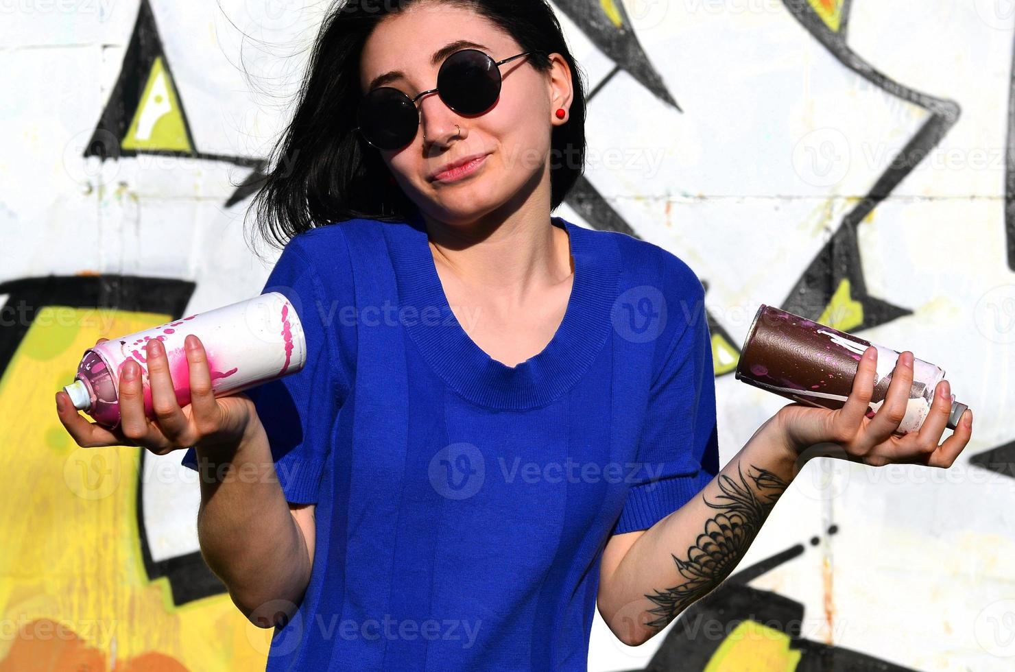 Porträt eines emotionalen jungen Mädchens mit schwarzen Haaren und Piercings. Foto eines Mädchens mit Aerosol-Farbdosen in den Händen auf einem Graffiti-Wand-Hintergrund. das Konzept der Straßenkunst und die Verwendung von Aerosolfarben