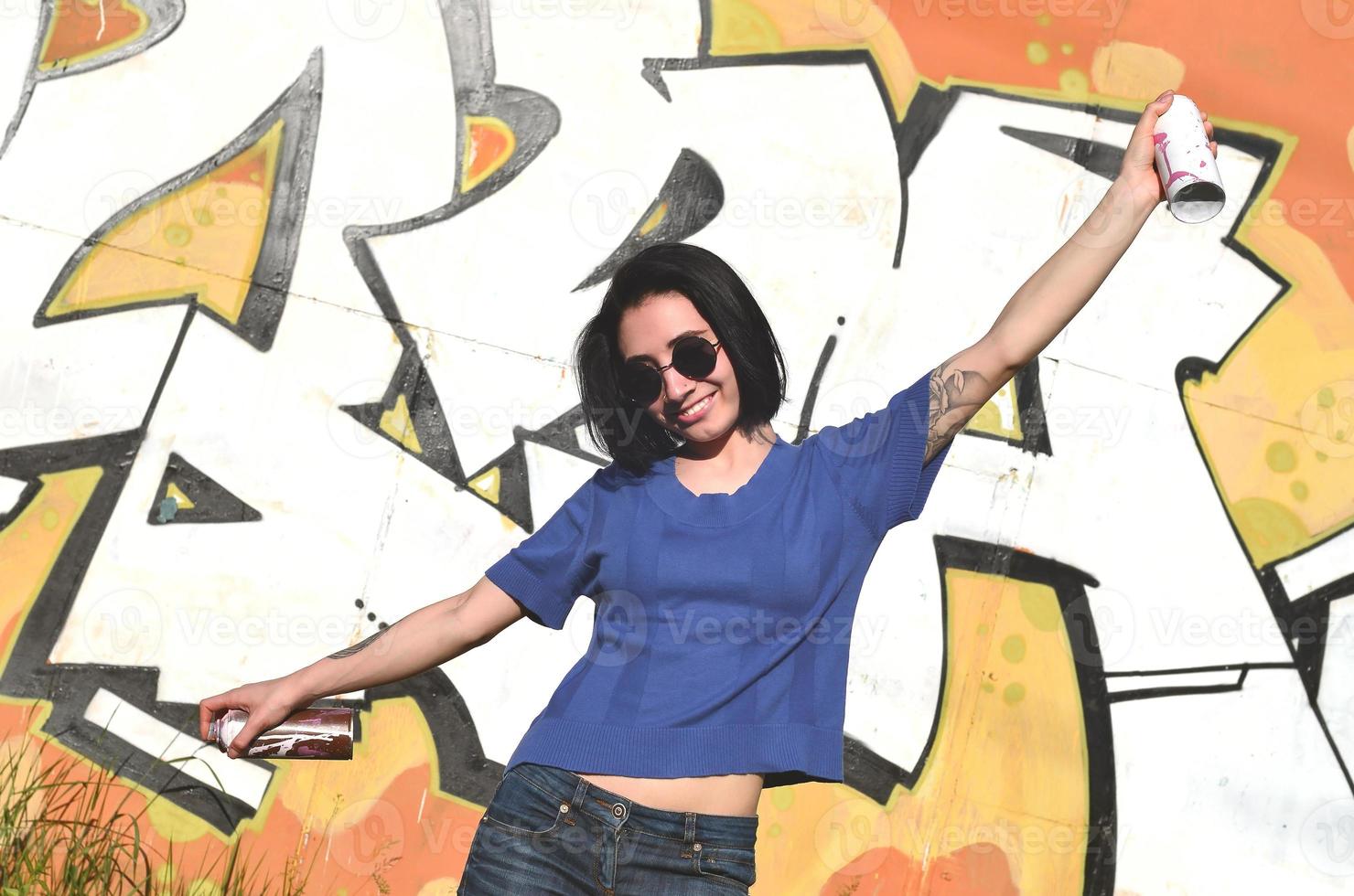 Porträt eines emotionalen jungen Mädchens mit schwarzen Haaren und Piercings. Foto eines Mädchens mit Aerosol-Farbdosen in den Händen auf einem Graffiti-Wand-Hintergrund. das Konzept der Straßenkunst und die Verwendung von Aerosolfarben