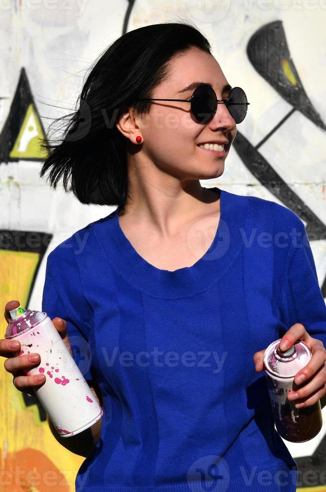 Porträt eines emotionalen jungen Mädchens mit schwarzen Haaren und Piercings. Foto eines Mädchens mit Aerosol-Farbdosen in den Händen auf einem Graffiti-Wand-Hintergrund. das Konzept der Straßenkunst und die Verwendung von Aerosolfarben