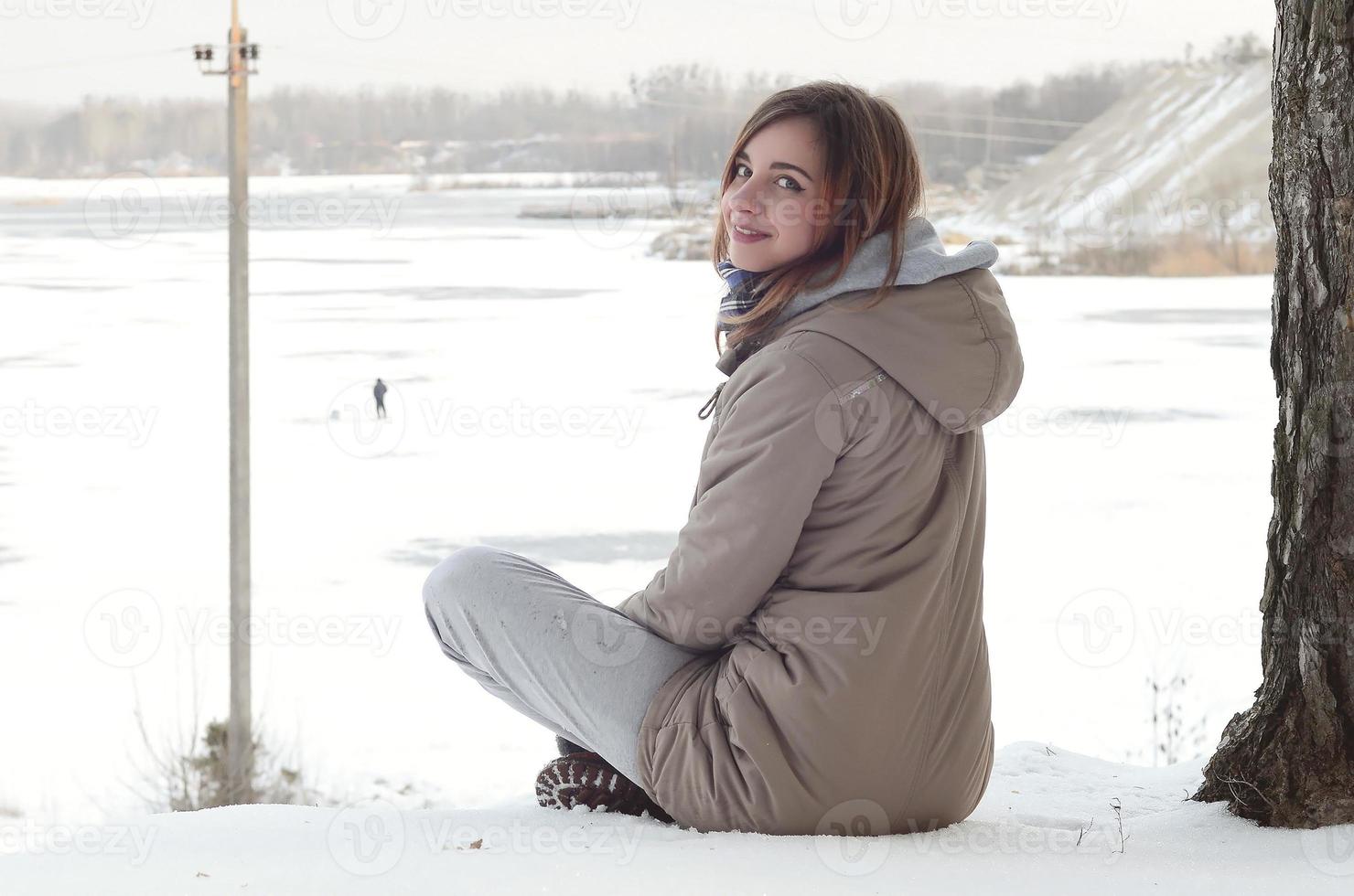 Ein junges kaukasisches Mädchen in einem braunen Mantel sitzt in der Nähe einer Klippe im Hintergrund einer Horizontlinie zwischen dem Himmel und einem zugefrorenen See im Winter foto