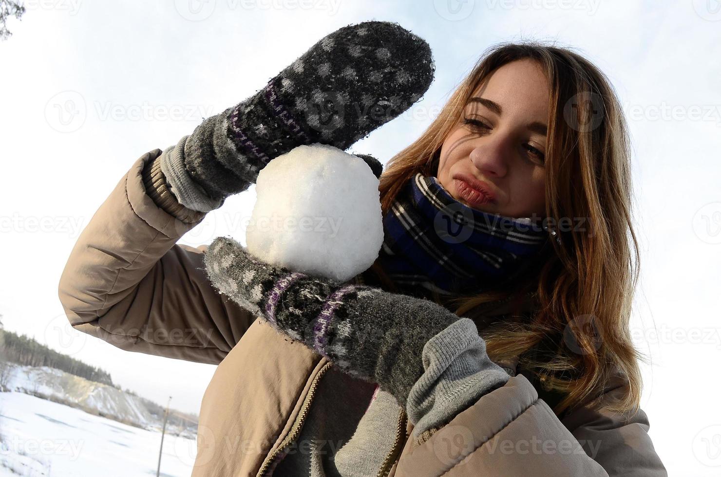 ein junges und fröhliches kaukasisches mädchen in einem braunen mantel hält im winter einen schneeball vor einer horizontlinie zwischen dem himmel und einem zugefrorenen see. Fisheye-Foto foto