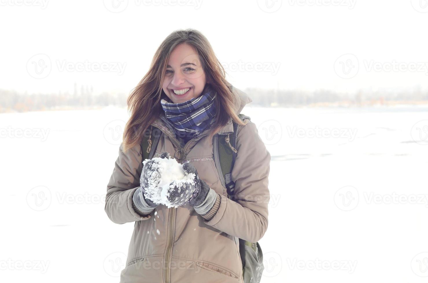 Ein junges und fröhliches kaukasisches Mädchen in einem braunen Mantel hält im Winter einen Schneeball im Hintergrund einer Horizontlinie zwischen dem Himmel und einem zugefrorenen See foto