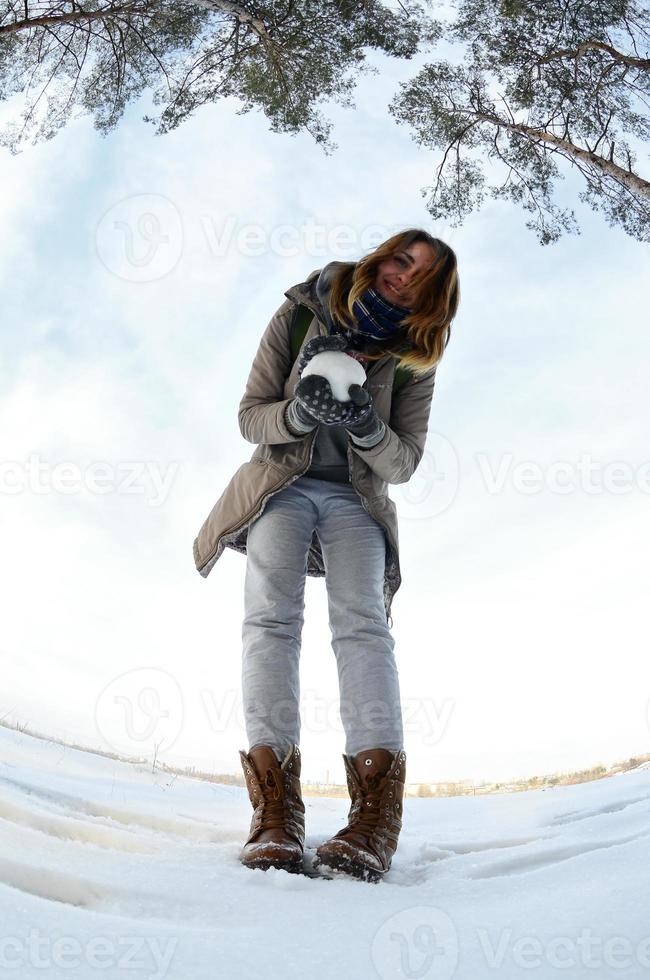 ein junges und fröhliches kaukasisches mädchen in einem braunen mantel hält im winter einen schneeball vor einer horizontlinie zwischen dem himmel und einem zugefrorenen see. Fisheye-Foto foto