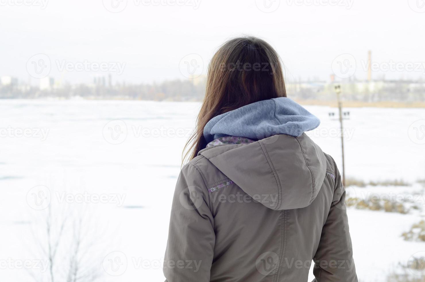 Ein junges kaukasisches Mädchen in einem braunen Mantel, das im Winter in die Ferne auf der Horizontlinie zwischen dem Himmel und dem zugefrorenen See starrt foto