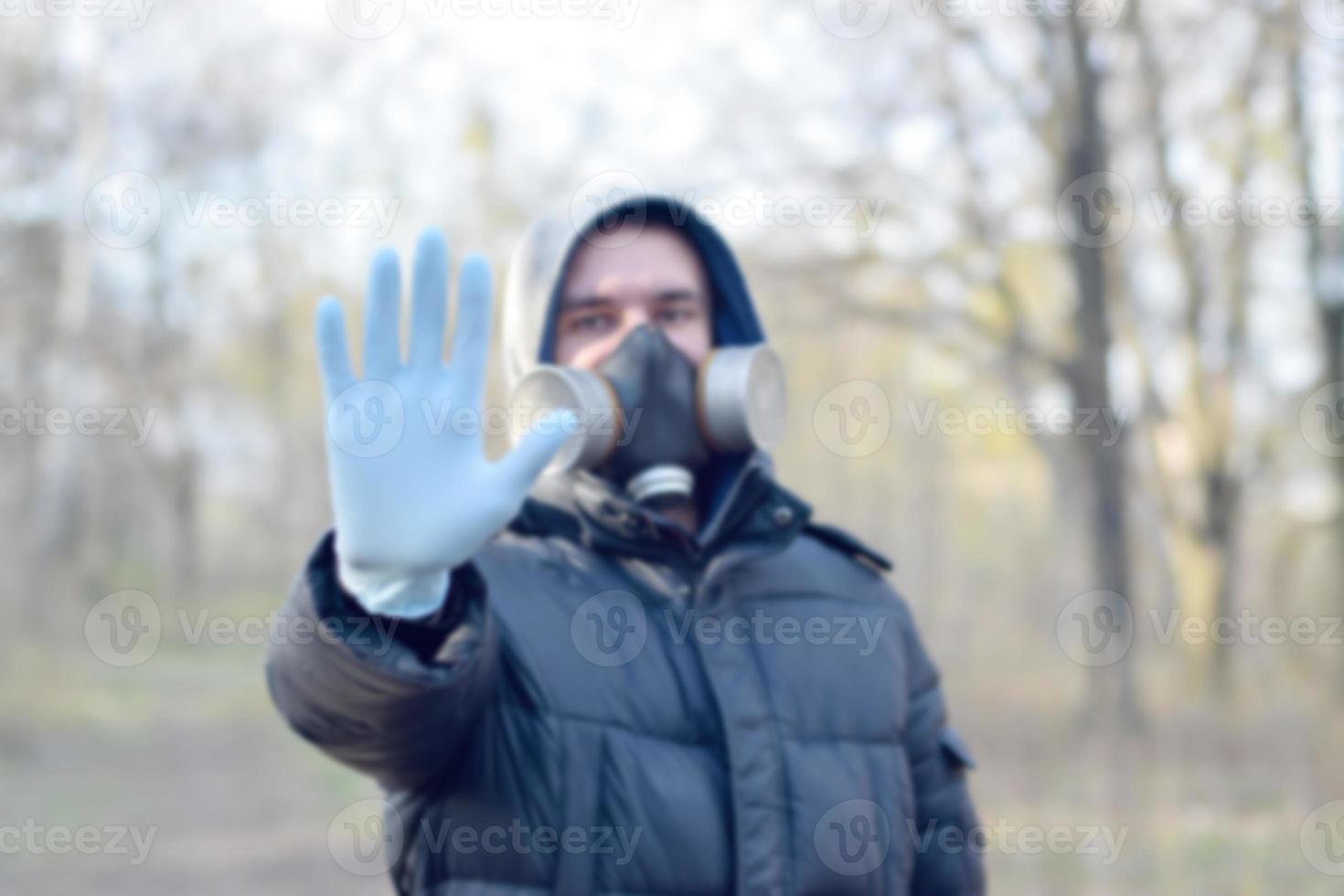 Verschwommenes Porträt eines jungen Mannes in Schutzgasmaske und Einweghandschuhen aus Gummi zeigt Stoppgeste im Freien im Frühlingsholz foto