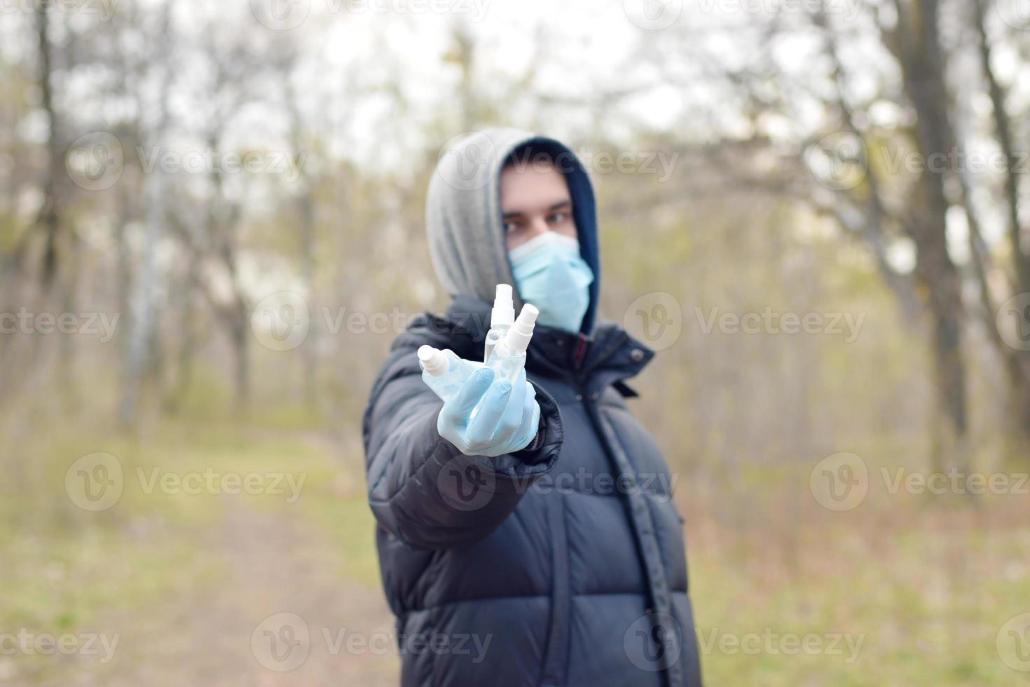Junger Mann in Schutzmaske zeigt Desinfektionssprühflaschen im Freien im Frühlingsholz foto