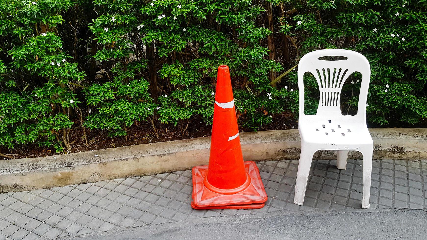 Orangefarbener Leitkegel und weißer Plastikstuhl befinden sich auf einem Fußweg mit weißer Blume und grüner Pflanze oder Baumhintergrund im Garten mit Kopierraum. Sitz mit Verkehrszeichenobjekt. foto