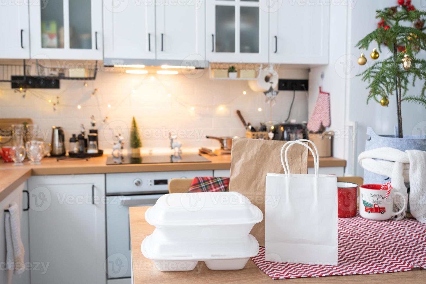 Lebensmittel-Lieferservice-Container auf dem Tisch Weiße Scandi-Festküche im Weihnachtsdekor. Silvesterabend, Zeitersparnis, zu faul zum Kochen, heiße Bestellung, Einweg-Plastikbox in Lichterkette. Attrappe, Lehrmodell, Simulation foto