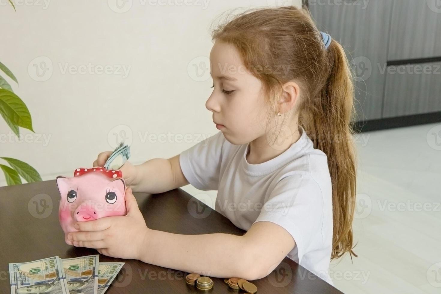 Finanzkompetenzkonzept, süßes kleines Mädchen, das konzentriert am Tisch sitzt, legt Banknoten in ein Sparschwein foto