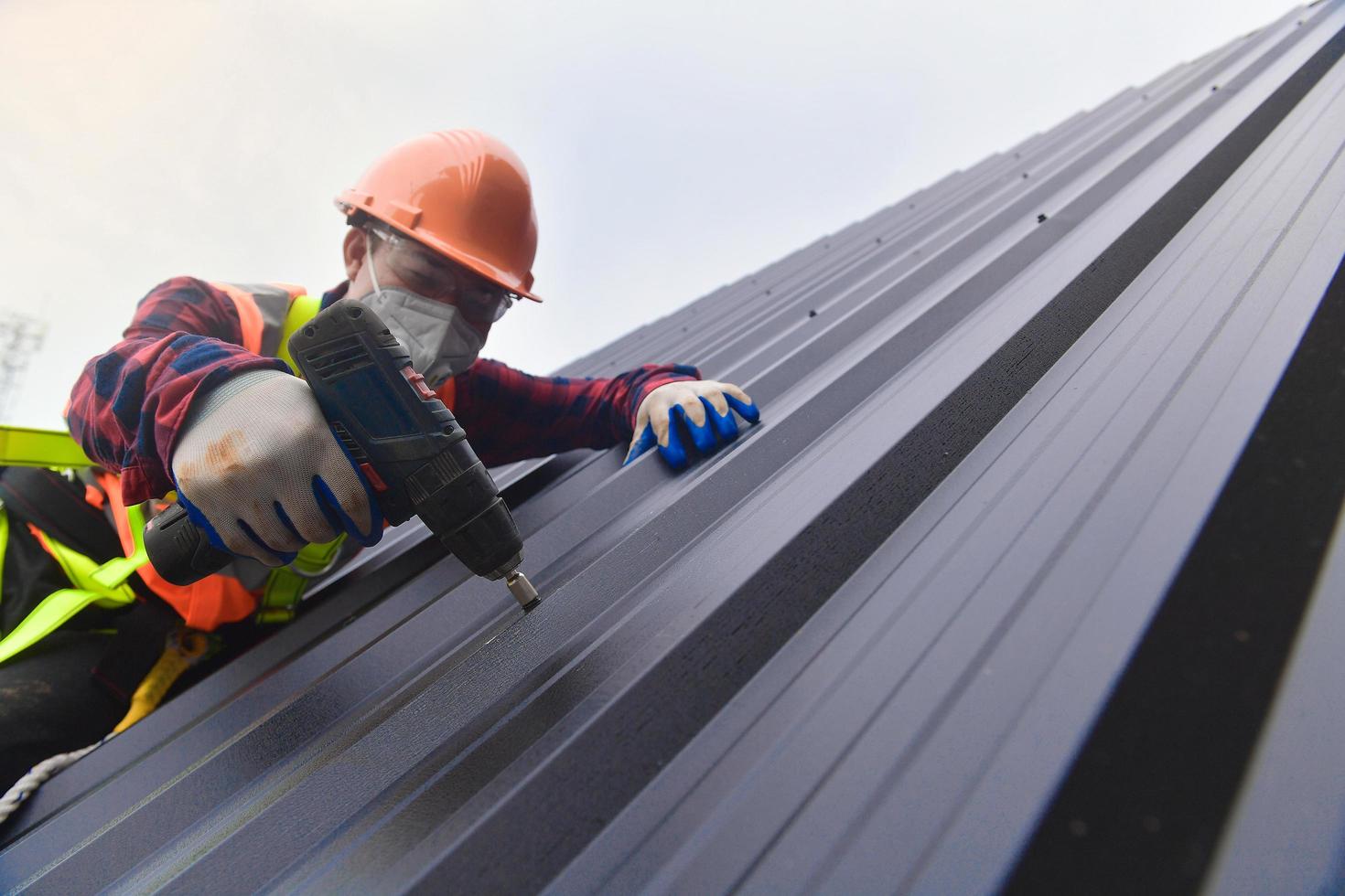 nahaufnahme bohrer dachdecker in schutzkleidung und handschuhen, dachdeckerwerkzeuge, installation neuer dächer im bau, elektrische bohrmaschine für neue dächer mit blech. foto