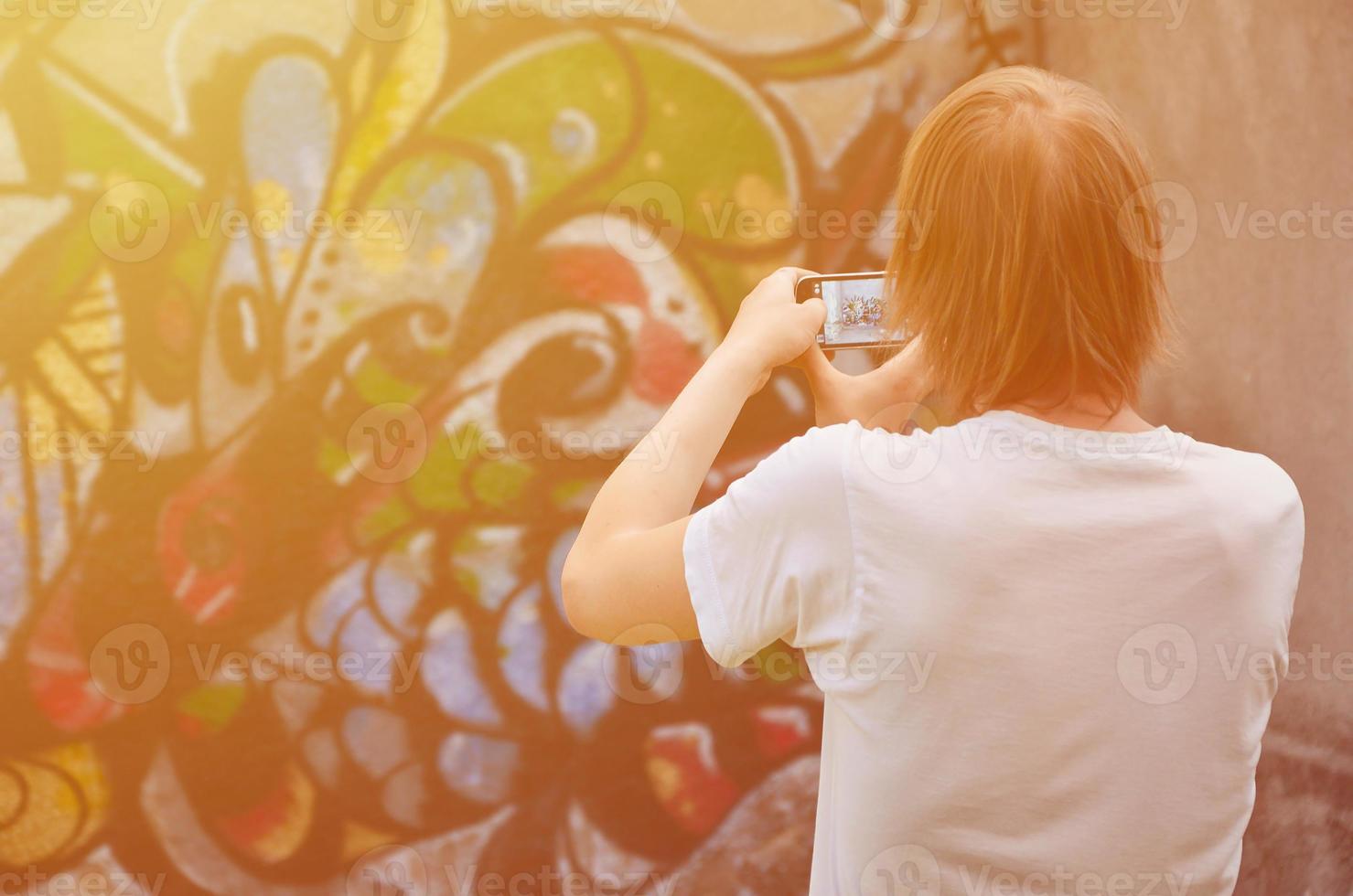 Foto beim Zeichnen von Graffiti auf einer alten Betonwand. ein junger, langhaariger blonder typ fotografiert seine fertige zeichnung auf einem smartphone. Straßenkunst- und Vandalismuskonzept