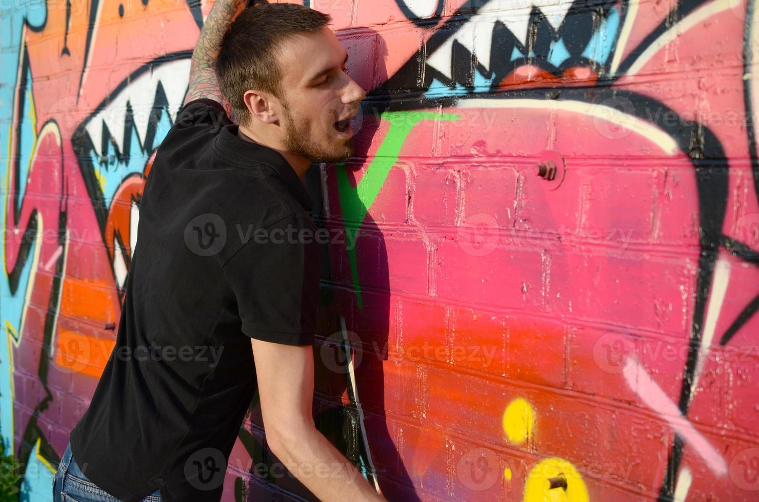 junger kaukasischer graffiti-künstler in schwarzem t-shirt mit silberner spraydose in der nähe von bunten graffiti in rosa tönen an der mauer. Street Art und zeitgenössischer Malprozess foto