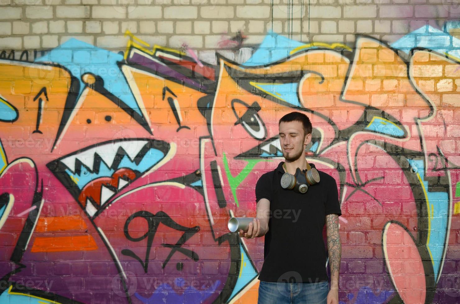 junger kaukasischer graffiti-künstler in schwarzem t-shirt mit silberner spraydose in der nähe von bunten graffiti in rosa tönen an der mauer. Street Art und zeitgenössischer Malprozess foto