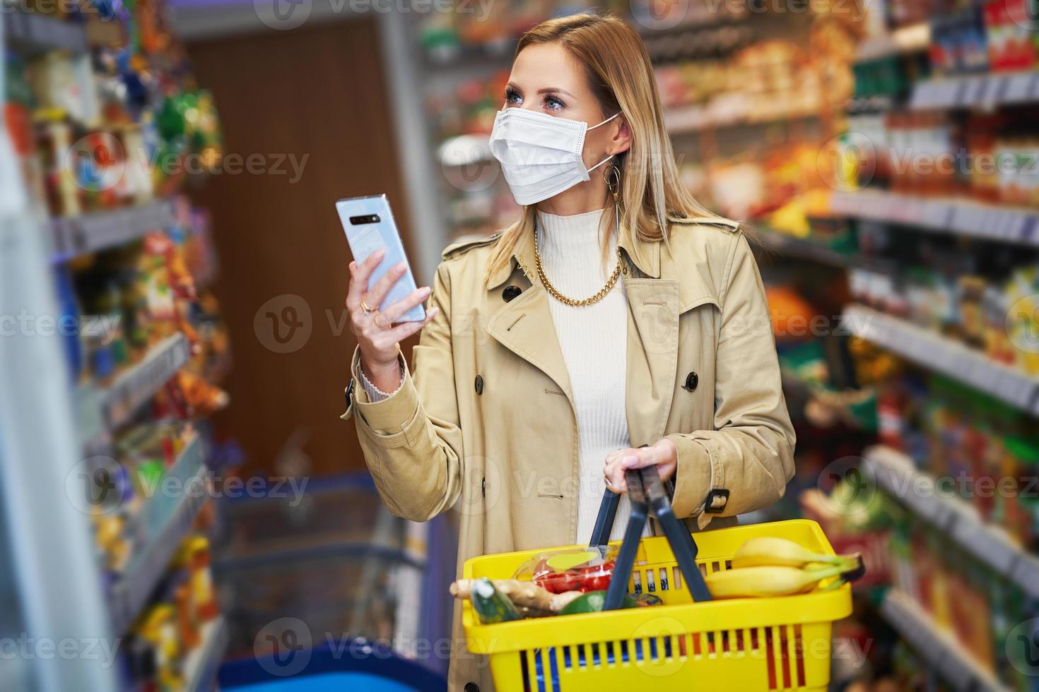 erwachsene frau in medizinischer maske, die smartphone verwendet und lebensmittel einkauft foto