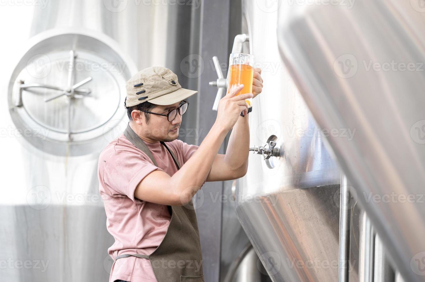 junger asiatischer arbeiter, der die brauereiqualität mit einem glas craft beer inspiziert und das visuelle erscheinungsbild nach der vorbereitung bewertet, während er in einer handwerklichen brauerei arbeitet. foto