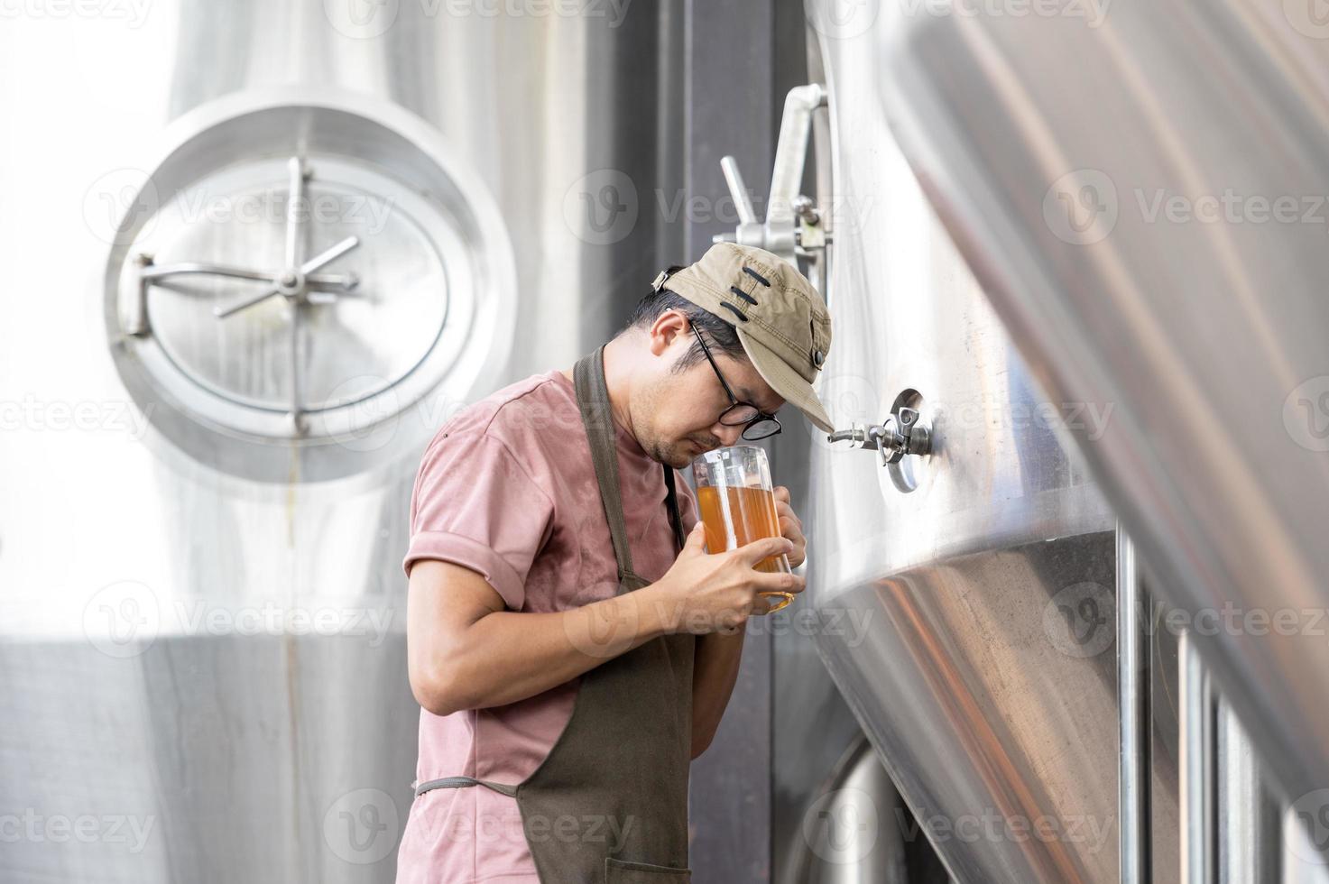 junger asiatischer arbeiter, der die brauereiqualität mit einem glas craft beer inspiziert und das visuelle erscheinungsbild nach der vorbereitung bewertet, während er in einer handwerklichen brauerei arbeitet. foto