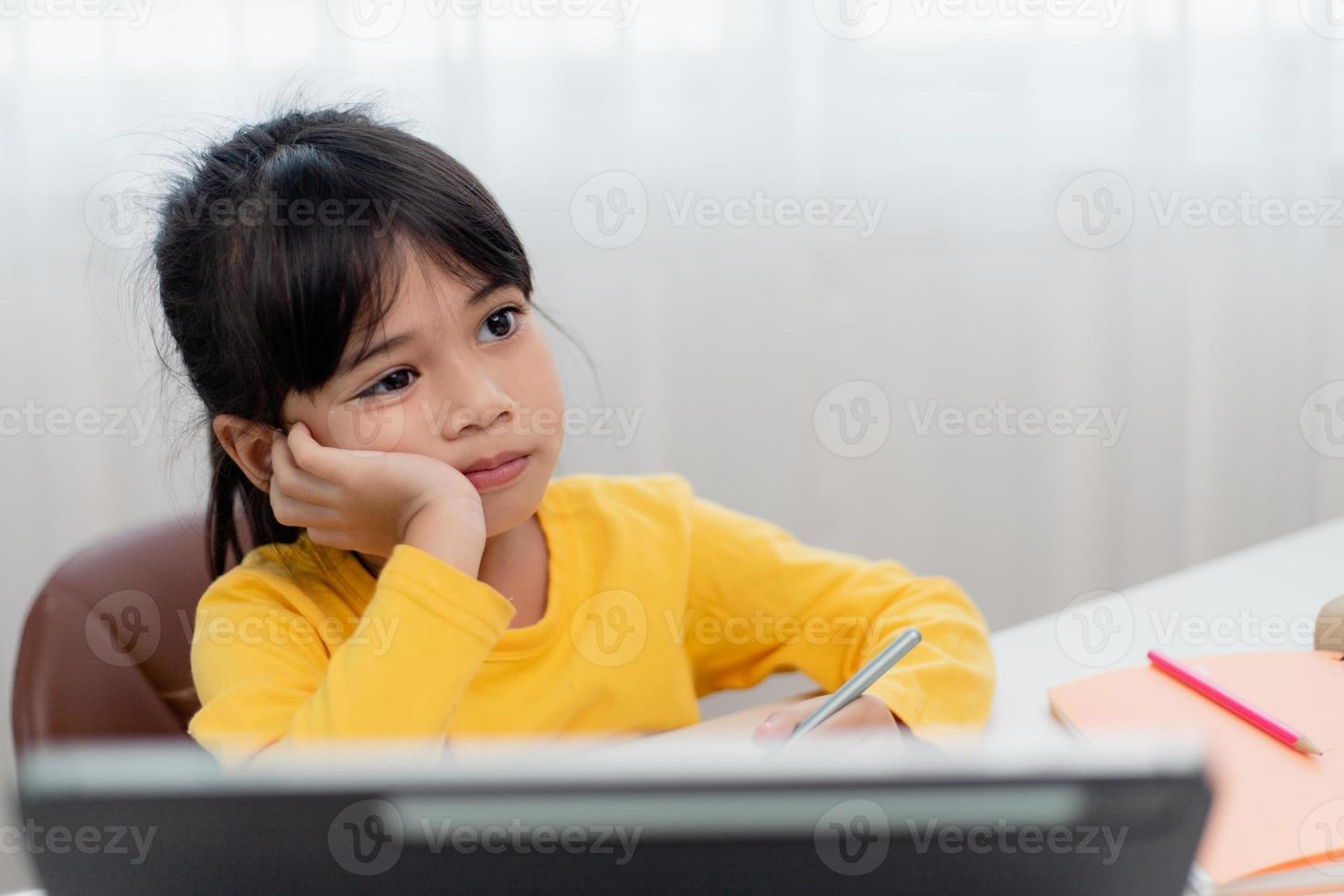 Kleines asiatisches Mädchen, das allein sitzt und mit gelangweiltem Gesicht herausschaut, Vorschulkind, das den Kopf auf den Tisch legt, mit trauriger Langeweile bei den Hausaufgaben, verwöhntes Kind foto