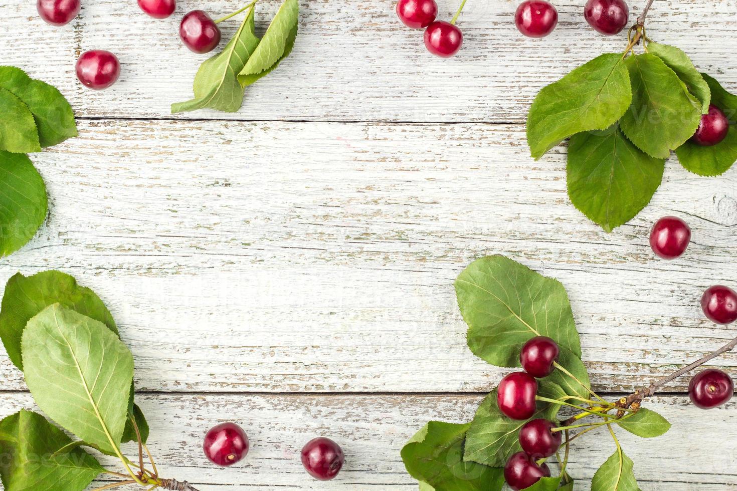 Rahmen aus roten reifen Kirschbeeren und grünen Blättern auf weißem Holzhintergrund. Ansicht von oben foto