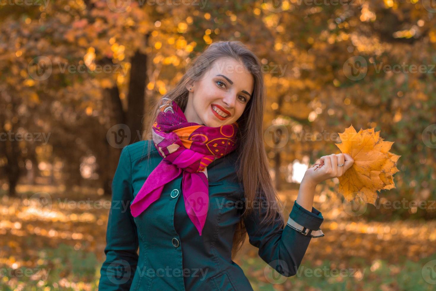 süßes junges mädchen in einem rosa schal um den hals steht im herbstpark und hält die blätter in der hand foto