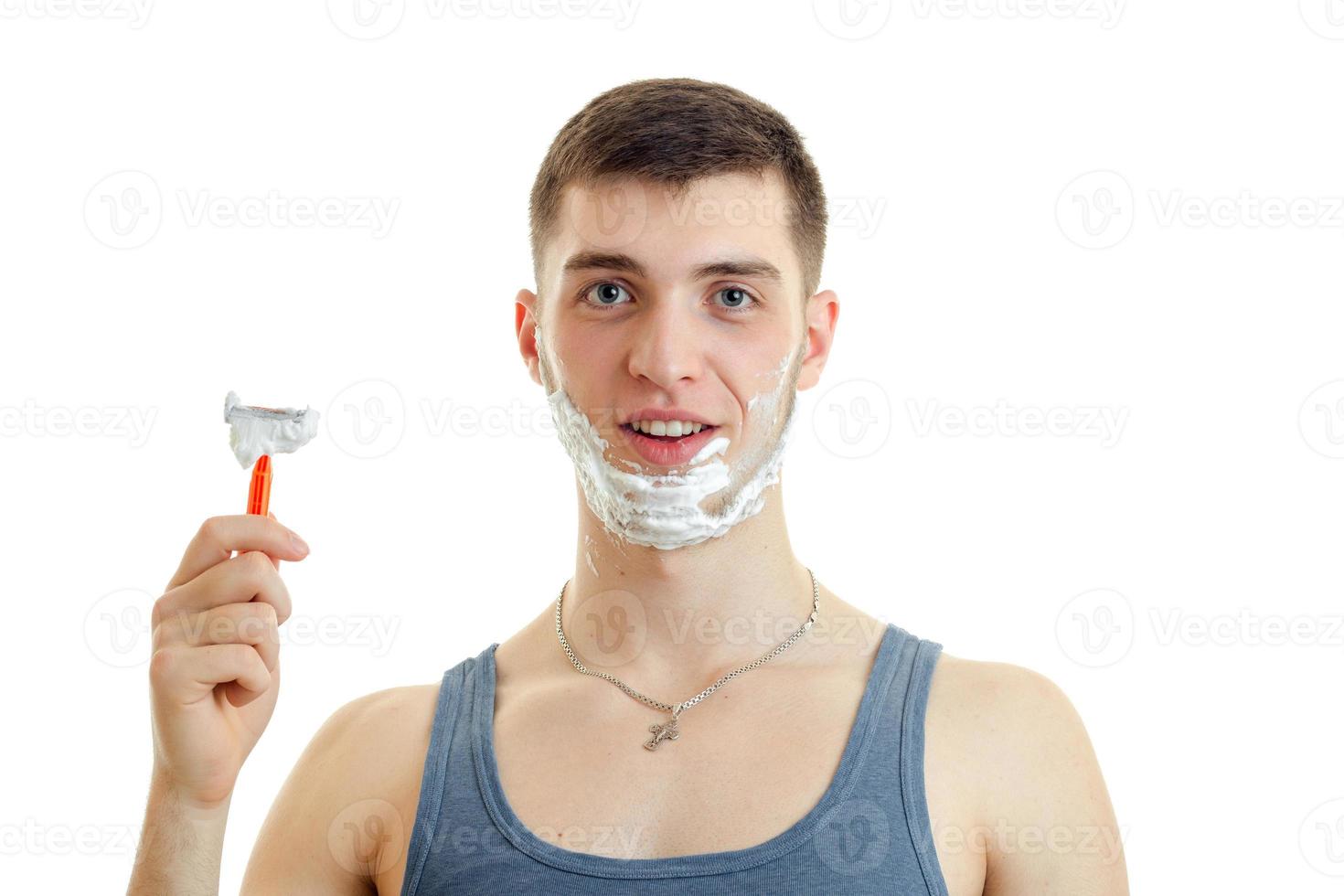 Horizontales Porträt eines jungen lächelnden Mannes, der mit Schaum auf seinem Gesicht steht und eine Rasiermaschine hält foto